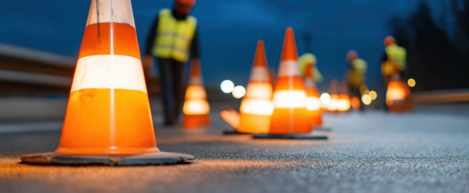 Lighted traffic cones are set out by workers in neon vests on a roadway.